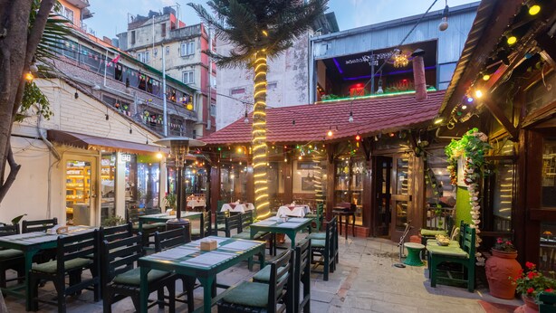 An outdoor dining area with tables and chairs, a palm tree, and a building with a red roof in the background. - Aawas Boutique Hotel (Kathmandu)