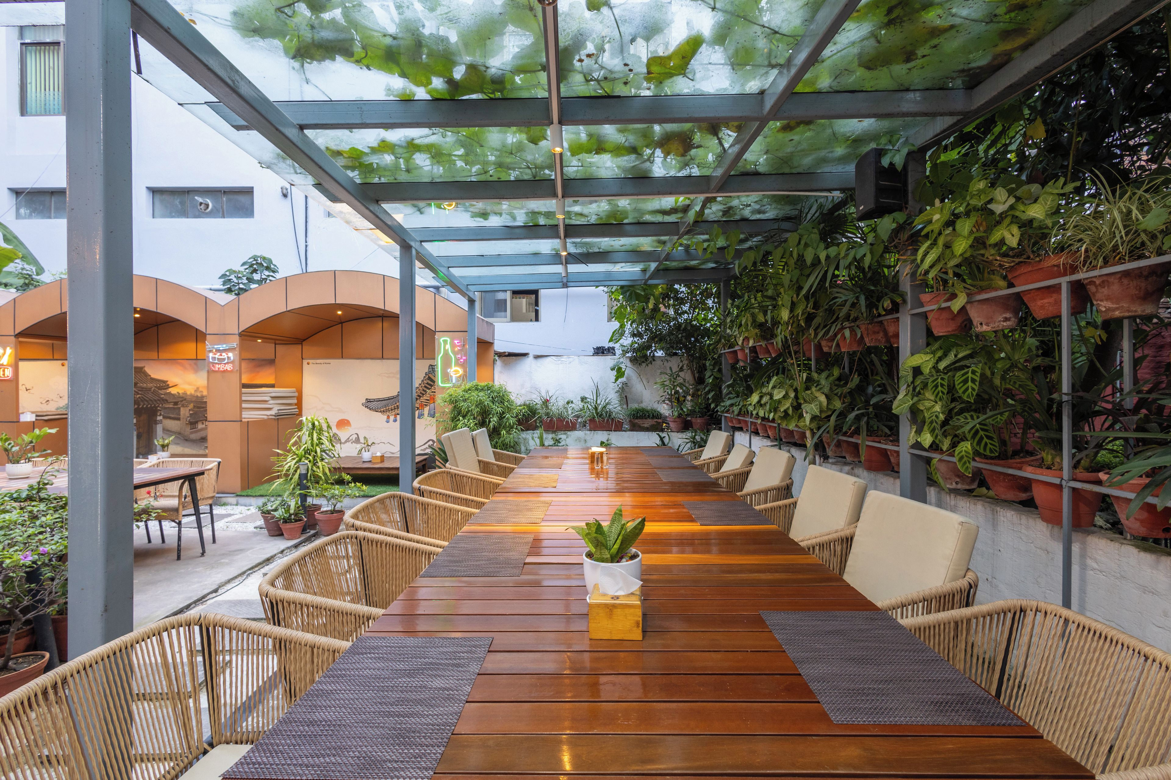 Un espace de restauration extérieur abrité, doté de tables en bois et de chaises en osier, entouré de plantes en pot et surmonté d’un plafond en verre.