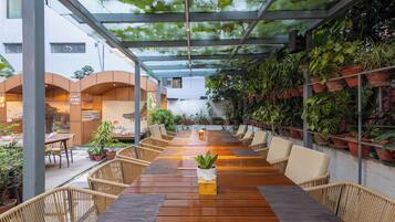 Un espacio exterior cubierto para comer con mesas de madera y sillas de mimbre, rodeado de plantas en macetas y un techo de vidrio.
