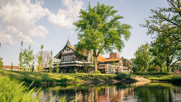 A traditional European-style building with a prominent tower, situated beside a calm water body, surrounded by lush greenery and other buildings. - Wapen van Hengelo Residence Suites - LOGIES ONLY - digital key by APP and email (Hengelo)