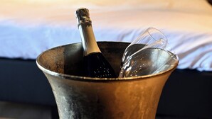 A metallic cup with a champagne bottle and bubbles, placed on a wooden table in front of a bed with a patterned headboard and pillows. - Wapen van Hengelo Residence Suites - LOGIES ONLY - digital key by APP and email (Hengelo)
