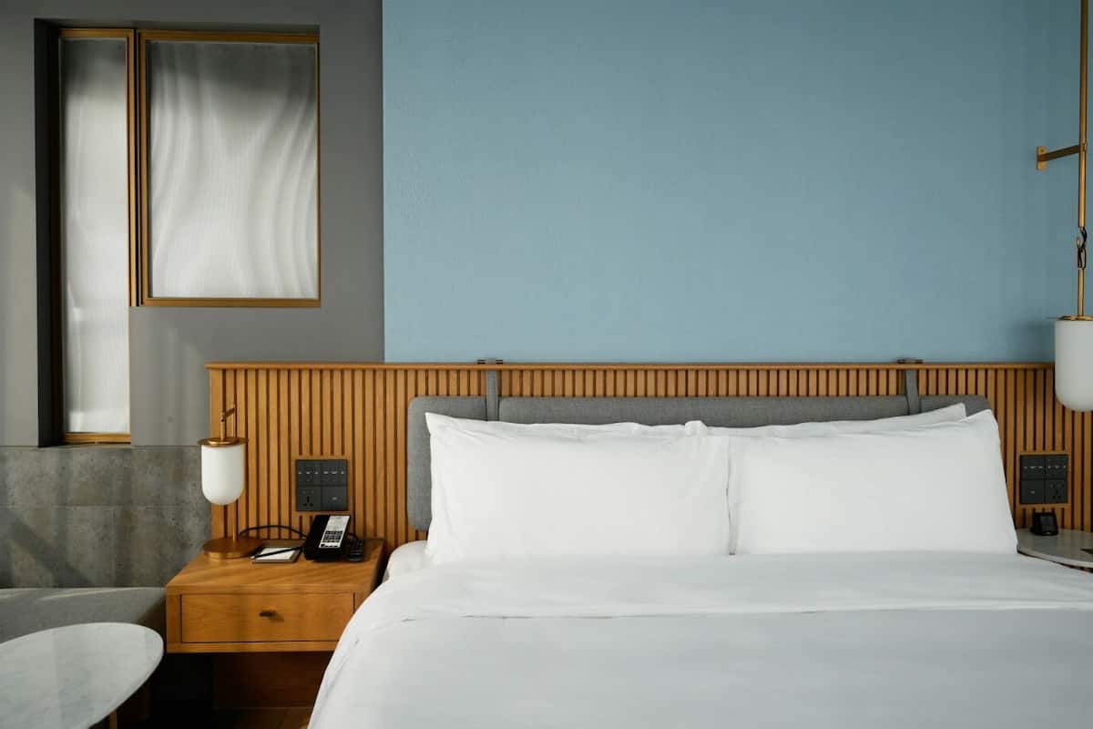 A hotel room with a wooden headboard, a bed with white linens, a nightstand with a lamp, a telephone, and a wall-mounted light fixture.