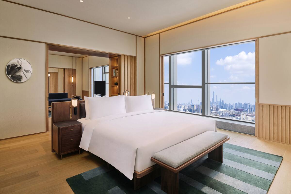 A modern hotel room with a large bed, a bench, a nightstand, a mirror, and a view of the city skyline.