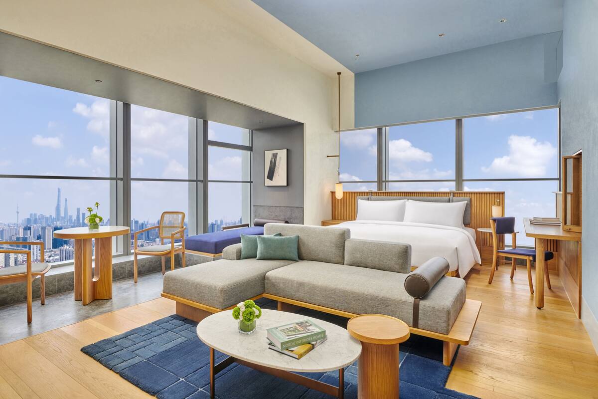 A modern hotel room with a large bed, a sofa, a dining area, and large windows offering a city view.