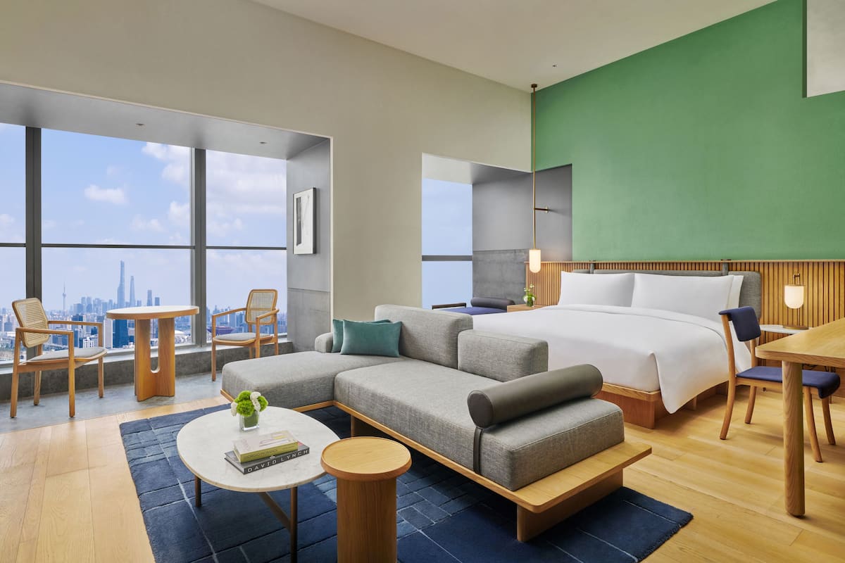 A modern hotel room with a sectional sofa, a bed, a dining table, and a city view.