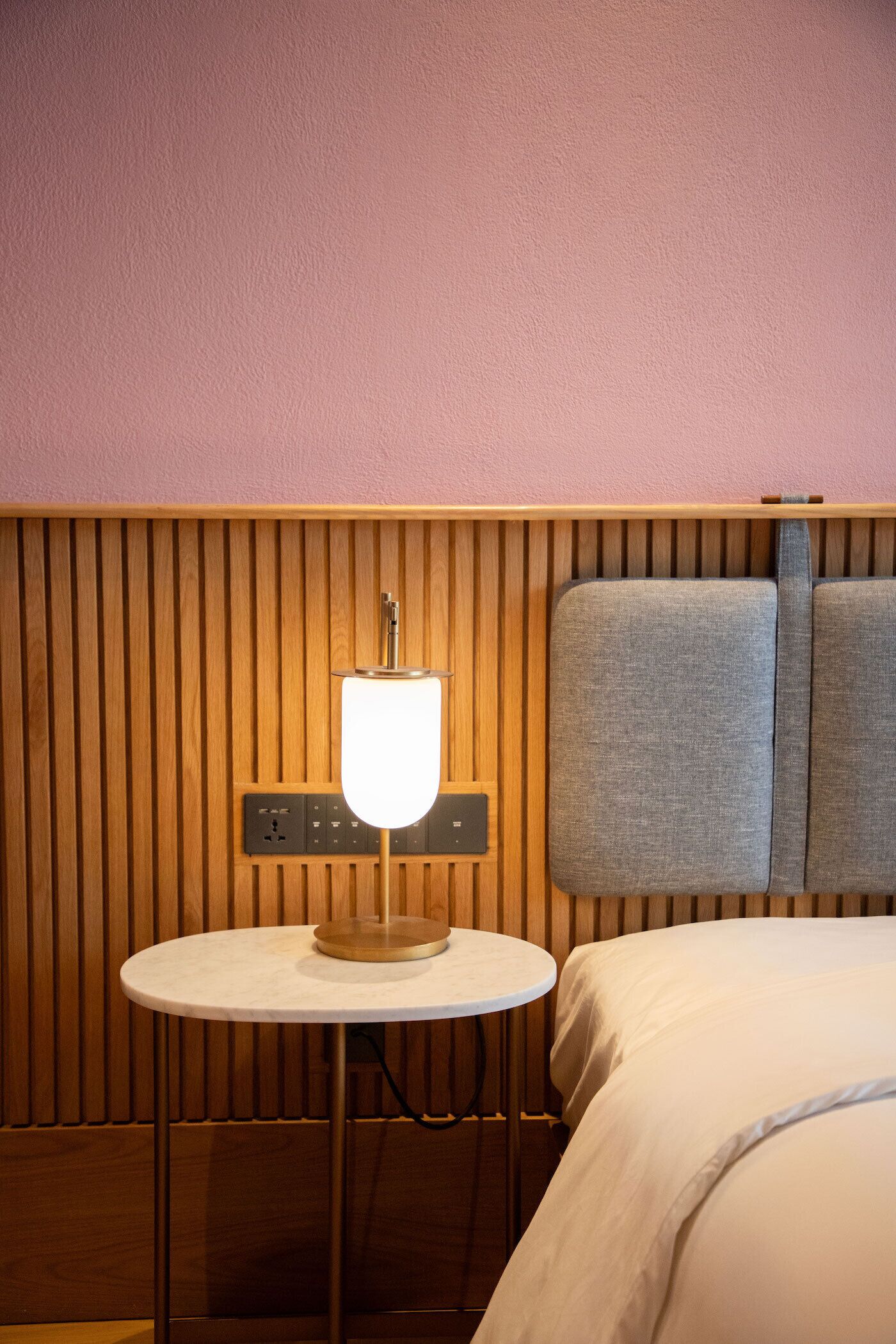 Hotellihuone, jossa on pyöreä pöytä, lamppu, sänky ja puupanelilla verhoiltu seinä.