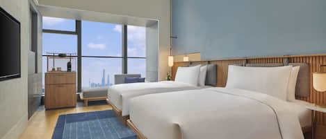 A modern hotel room with two beds, a large window offering a city view, a flat-screen TV, and a small desk area.