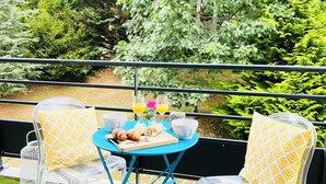 Un balcon avec une table bleue dressée pour le petit-déjeuner, deux chaises dotées de coussins à motifs jaunes, et une vue sur une végétation luxuriante.