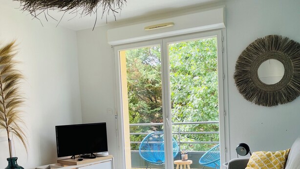 Un salon moderne avec un téléviseur à écran plat, un canapé gris agrémenté de coussins jaunes, un tapis tissé et une grande fenêtre donnant sur un paysage verdoyant, avec des chaises bleues à l’extérieur.
