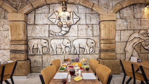 A dining area with a stone wall featuring animal carvings, a wooden table set with plates, glasses, and a variety of food items. - Legend Tulip Hotel (Antalya)