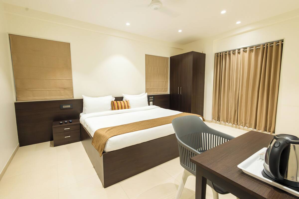 A modern hotel room with a large bed, a desk with a kettle, a chair, and a wardrobe.