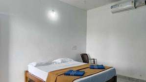 Ein Einzelbett mit Holzrahmen, weißer Bettwäsche und einer braunen Decke. Ein Stuhl und eine Klimaanlage befinden sich an der Wand.