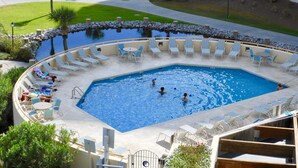 Una amplia piscina circular con tumbonas y una pequeña mesa, rodeada de jardines cuidados y un edificio al fondo.