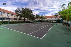 An outdoor tennis court with a green surface, surrounded by a fence, trees, and buildings with red roofs. - 4BR family retreat near Disney w/ patio (Kissimmee)
