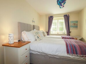 A bedroom with two beds, a bedside table with a lamp, a window with curtains, and a wall decoration. - Pen-y-les (Wadebridge)