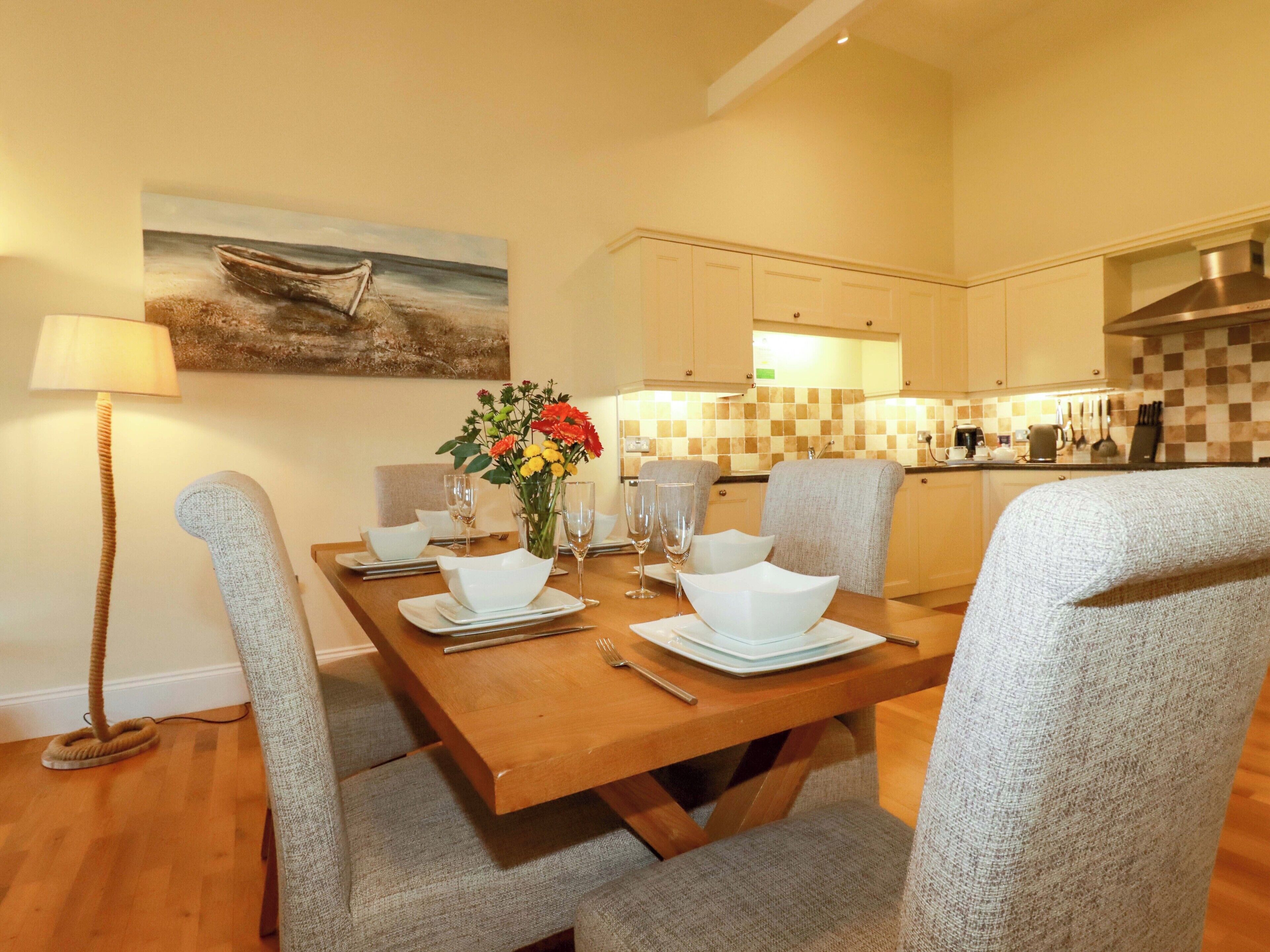 A dining area with a wooden table set for four, a vase of flowers, and a painting on the wall.