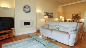 A living room with a sofa, a glass coffee table, a flat-screen TV, a fireplace, and a kitchen in the background. - Pen-y-les (Wadebridge)