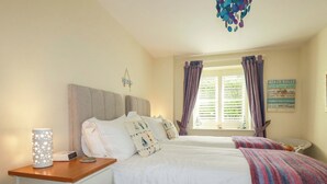 A bedroom with two beds, a bedside table with a lamp, a window with curtains, and a wall decoration. - Pen-y-les (Wadebridge)