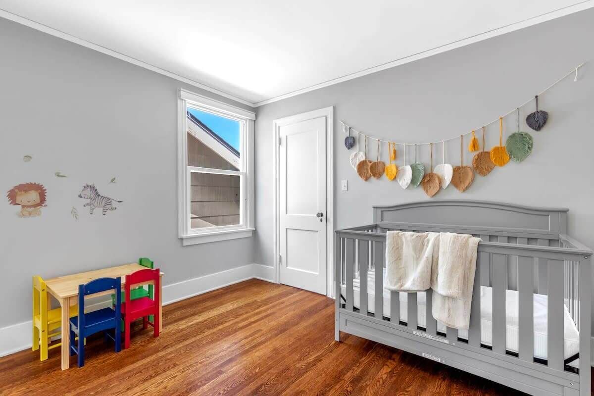 Ein Zimmer mit Holzboden, einem Babybett, einem Schreibtisch mit Stühlen und einem Fenster.