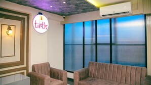 A hotel lobby with a reception desk, two sofas, a neon sign, and large windows with a view. - Hotel Tirth by Sonu Cafe (Ujjain)