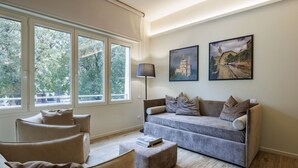 Ein modernes Wohnzimmer mit einem grauen Sofa, einem kleinen runden Tisch und einer Lampe. Große Fenster bieten Blick auf Bäume und ein Gebäude.