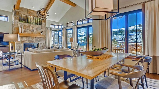 Un salon spacieux avec une table à manger en bois, des sièges confortables, une cheminée et de grandes fenêtres offrant une vue sur les montagnes.