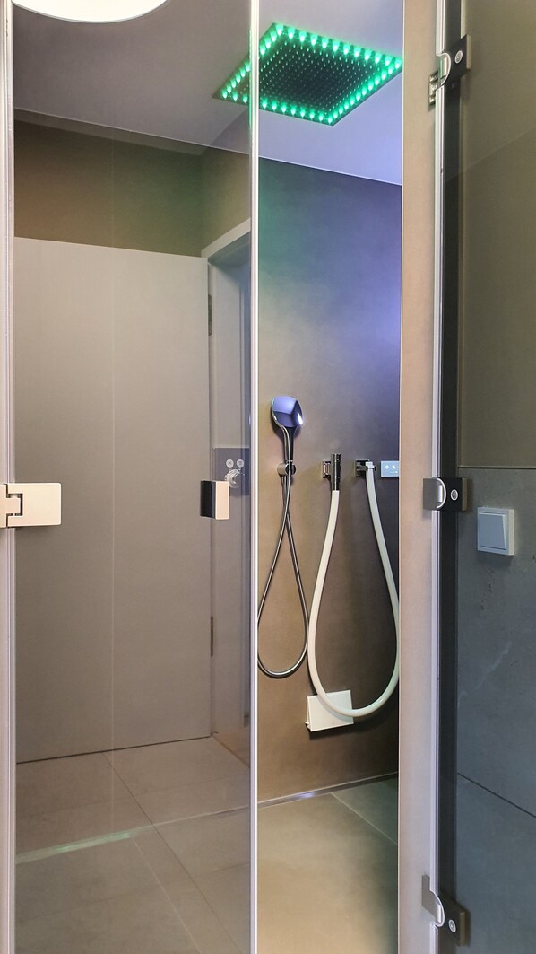 Un baño moderno con mampara de vidrio, ducha con cabezal de ducha manual y luz de techo LED verde.