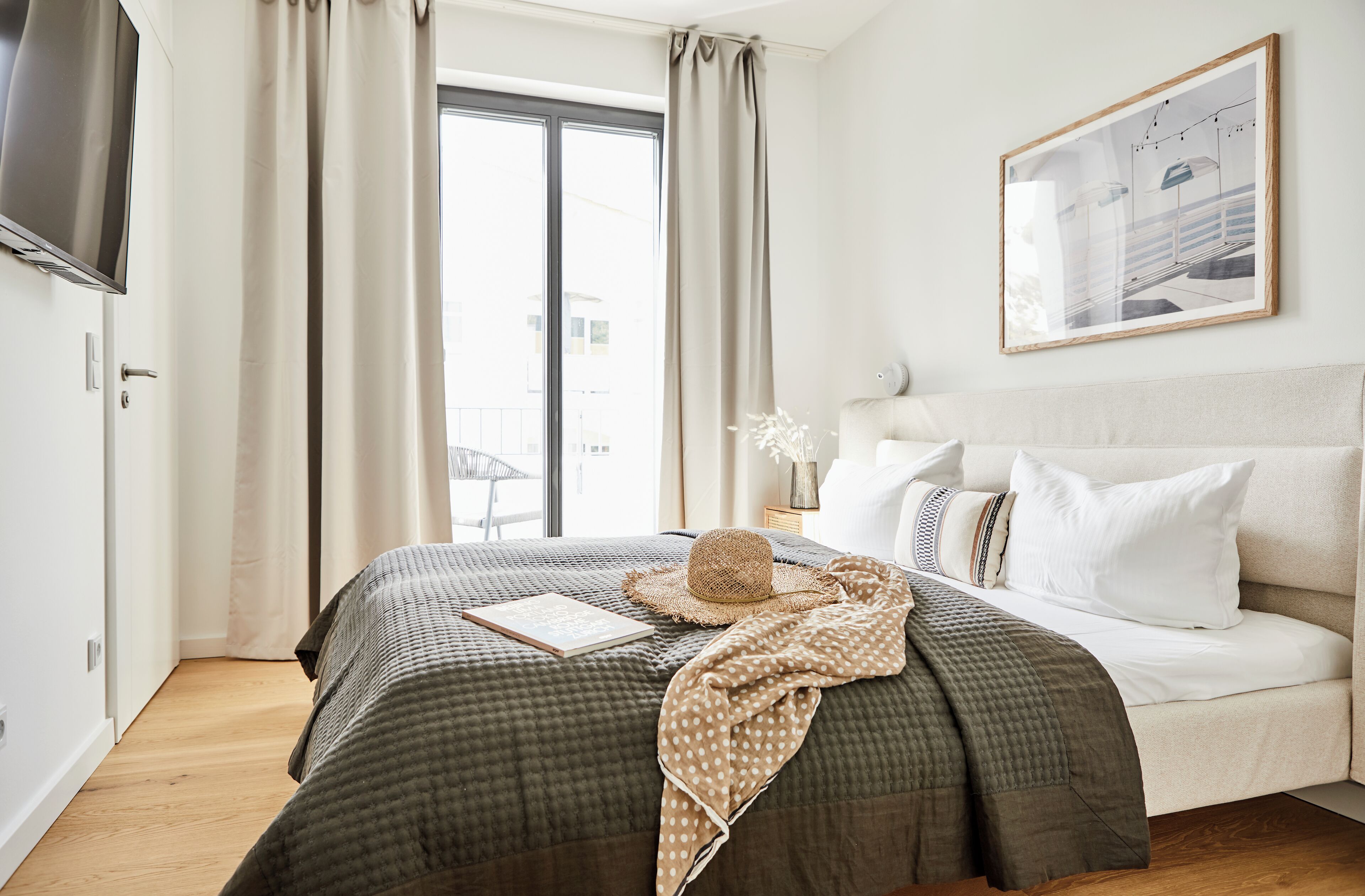 Un dormitorio con cama, televisor, ventana con cortinas, un cuadro enmarcado en la pared y una mesita de noche con lámpara y un florero.