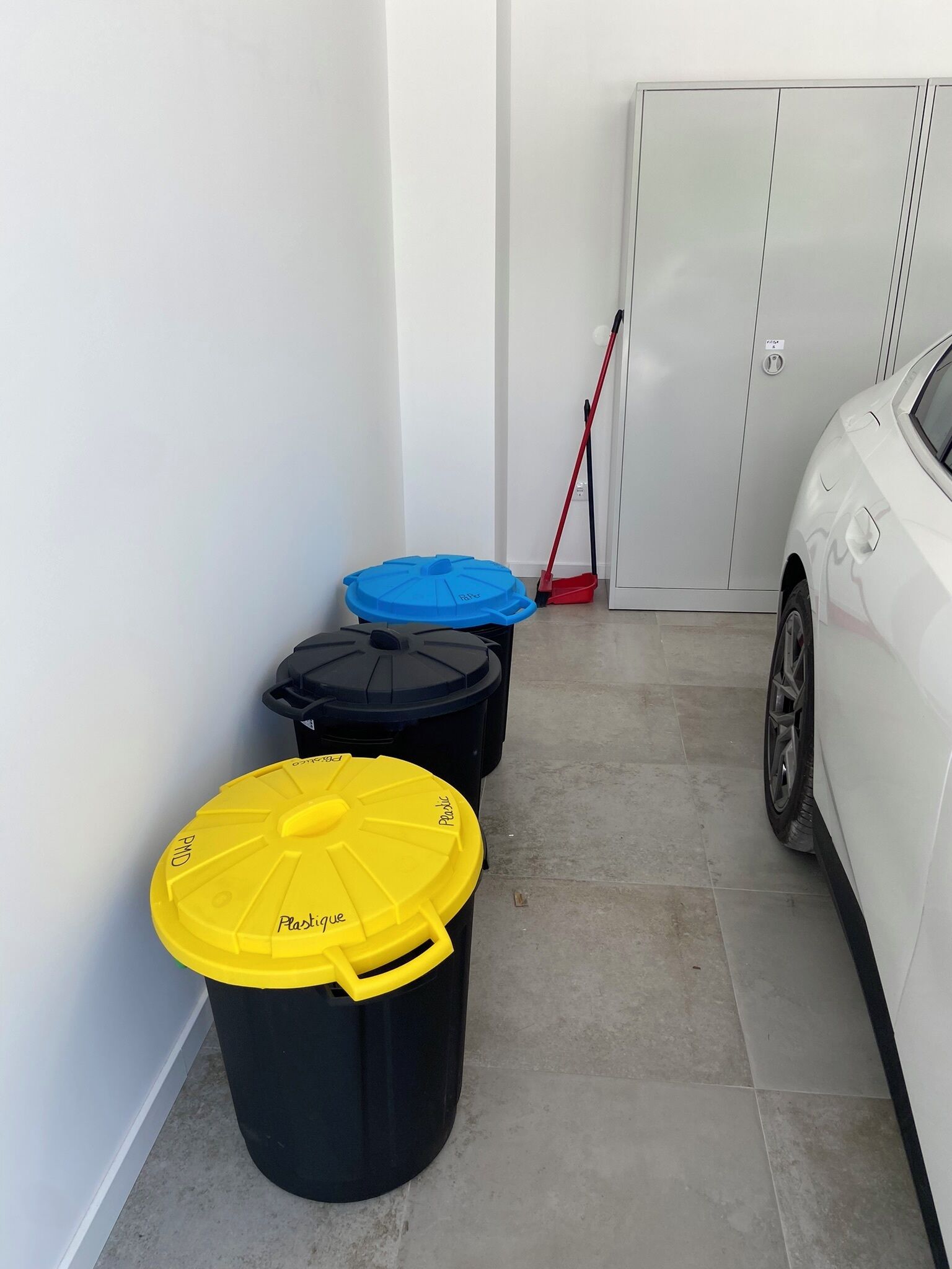 Un garage contient trois poubelles étiquetées « Plastique », « Papier » et « Déchets ». Un balai rouge est appuyé contre un mur, et une voiture blanche est garée à droite.
