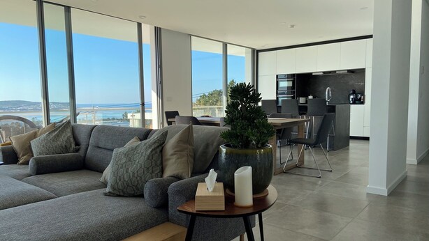 A modern living room with a large gray sectional sofa, a small wooden table with a vase and candles, and a view of the sea through large windows.