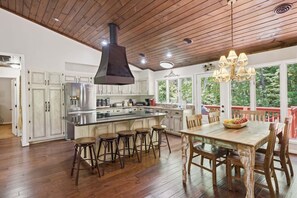 A spacious kitchen with a large island, wooden flooring, and a view of the outdoors through large windows.
