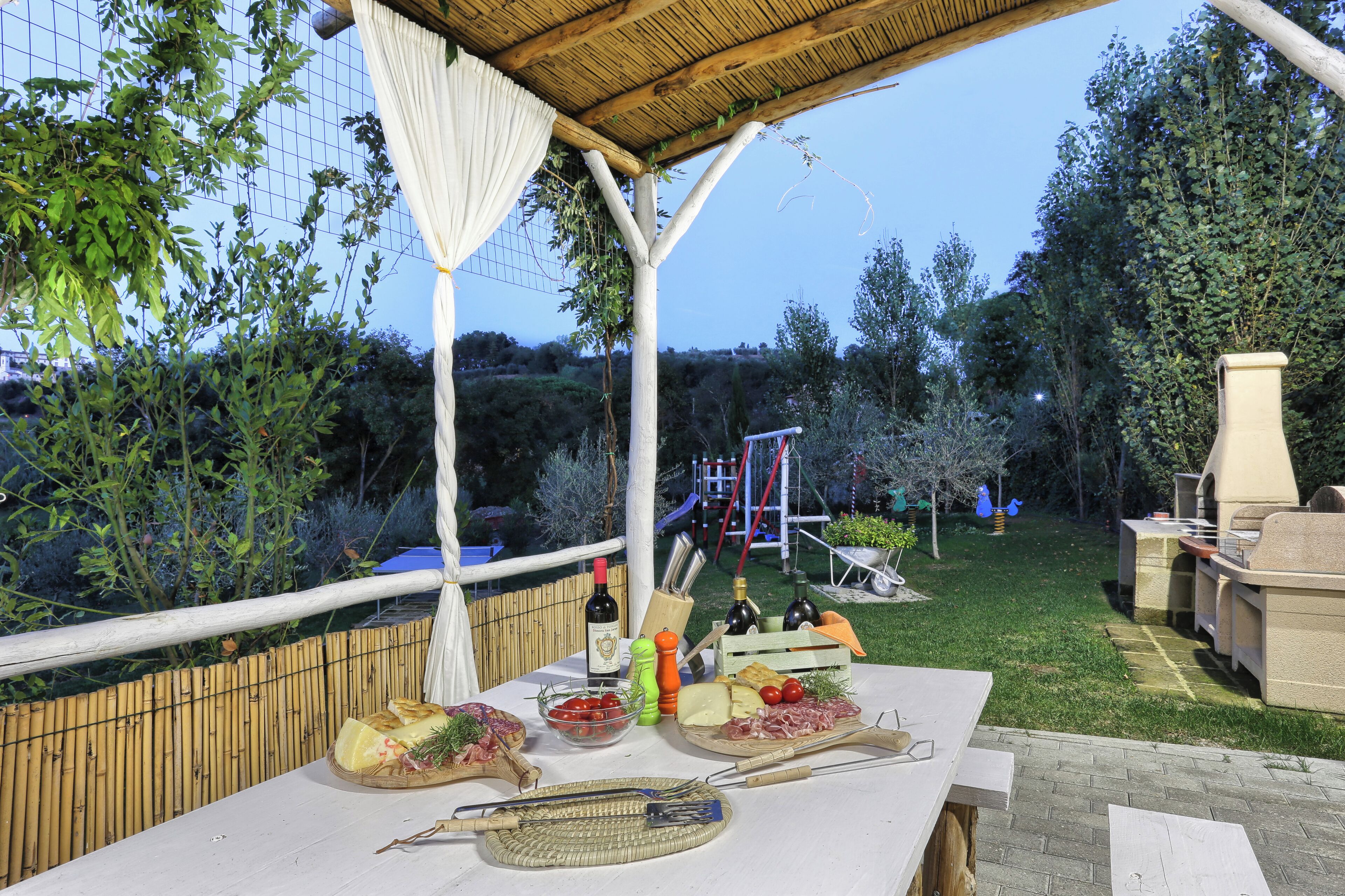 An outdoor dining area with a table set for a meal, surrounded by greenery and a barbecue grill.
