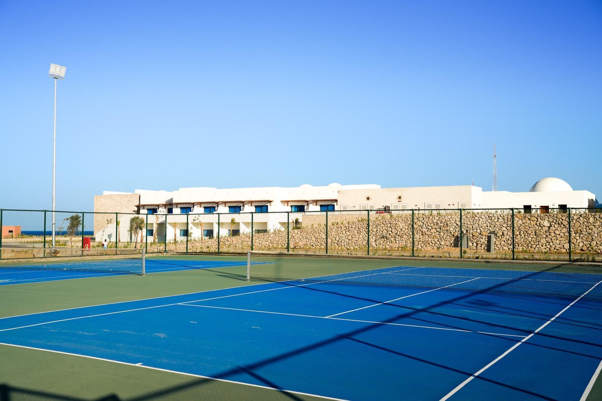 Una cancha de tenis con red, rodeada por un muro de piedra y un edificio con múltiples ventanas.