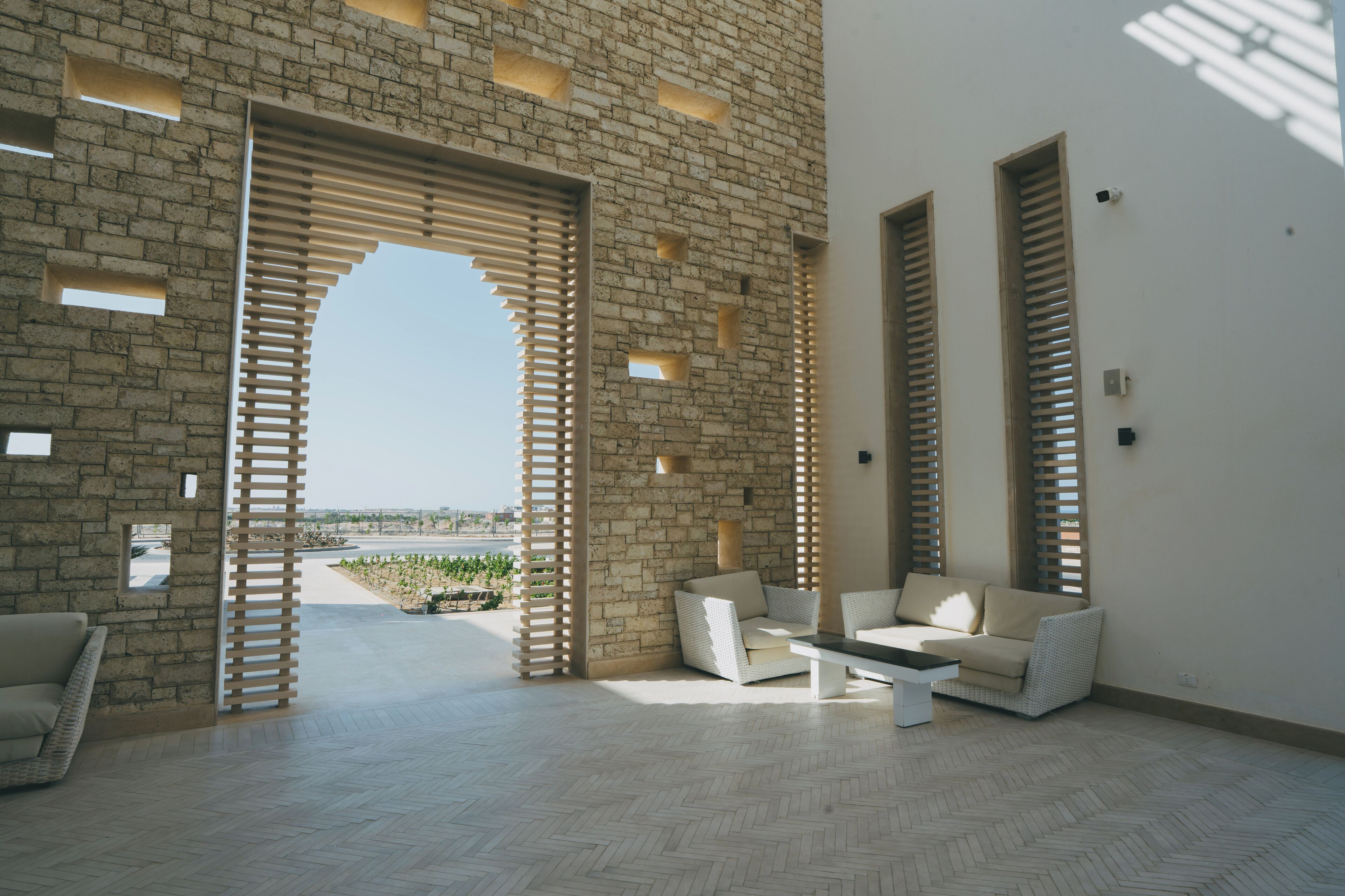 A spacious room with a herringbone floor, a white sofa set, and a coffee table. There are large windows with wooden shutters and a view of the outdoors.
