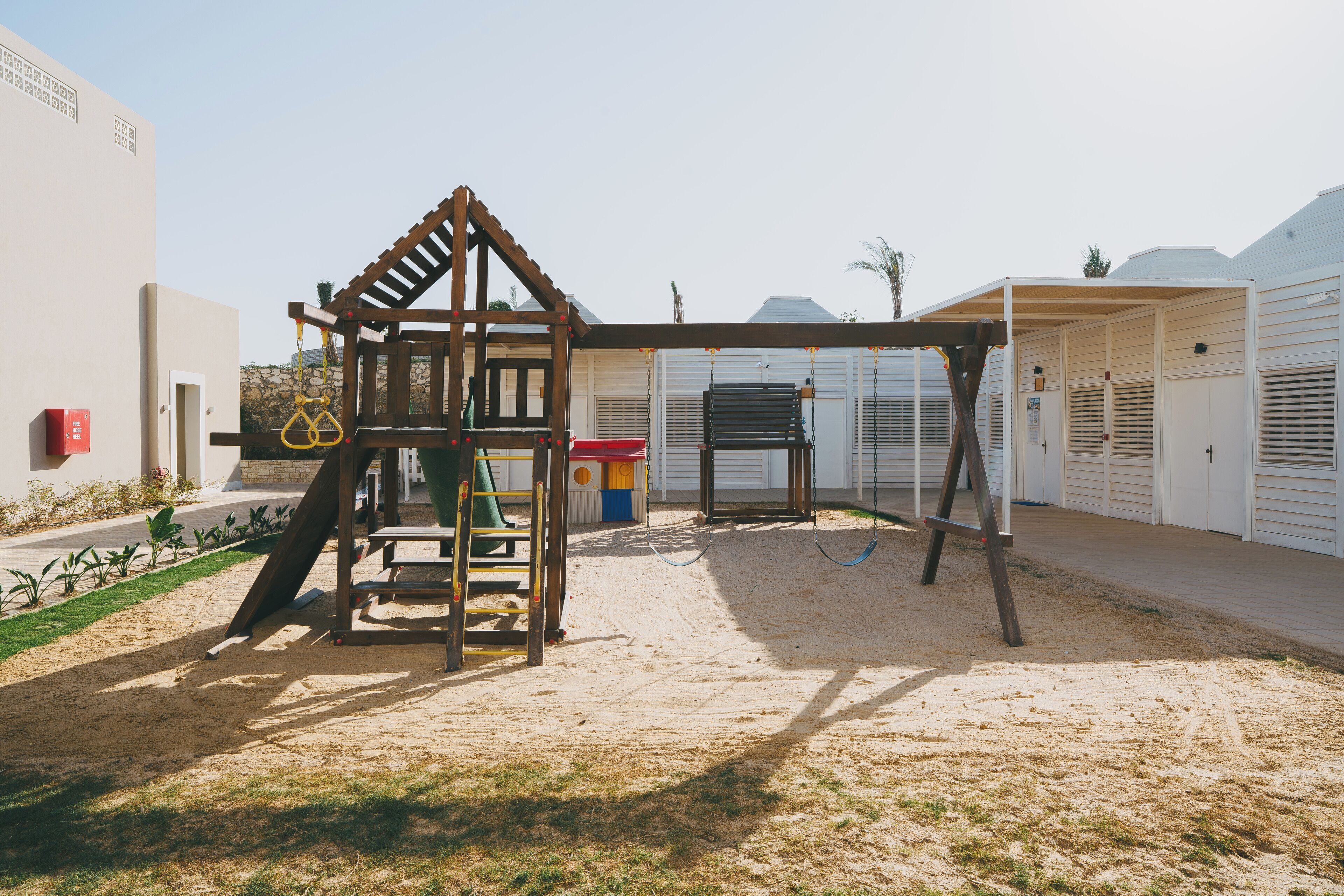 A playground with a wooden swing set and slide, surrounded by a sandy area with a building in the background.
