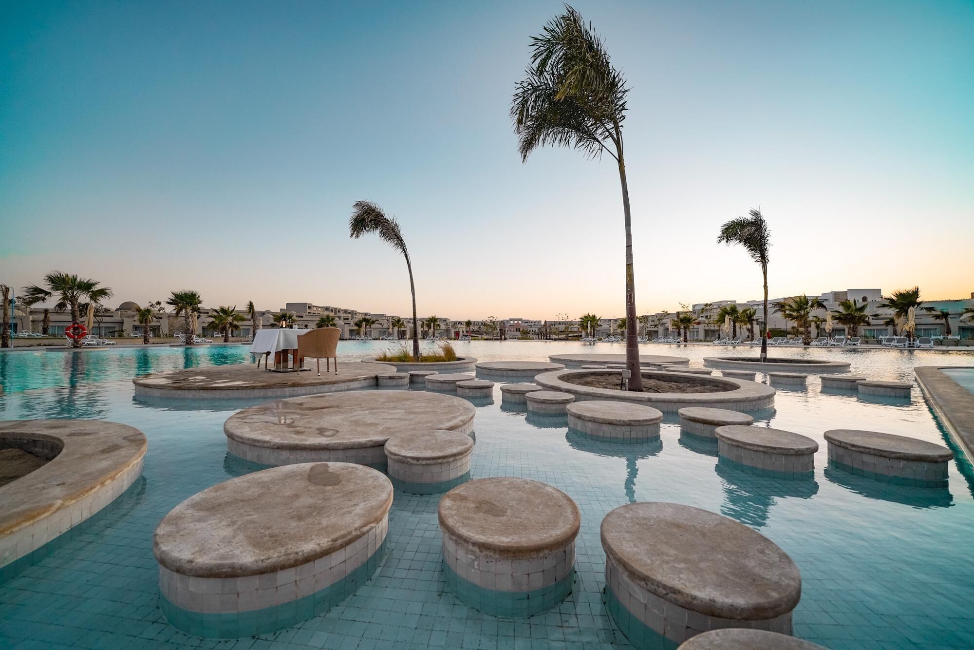 Zona de piscina con asientos circulares y palmeras, rodeada de edificios y un cielo despejado.