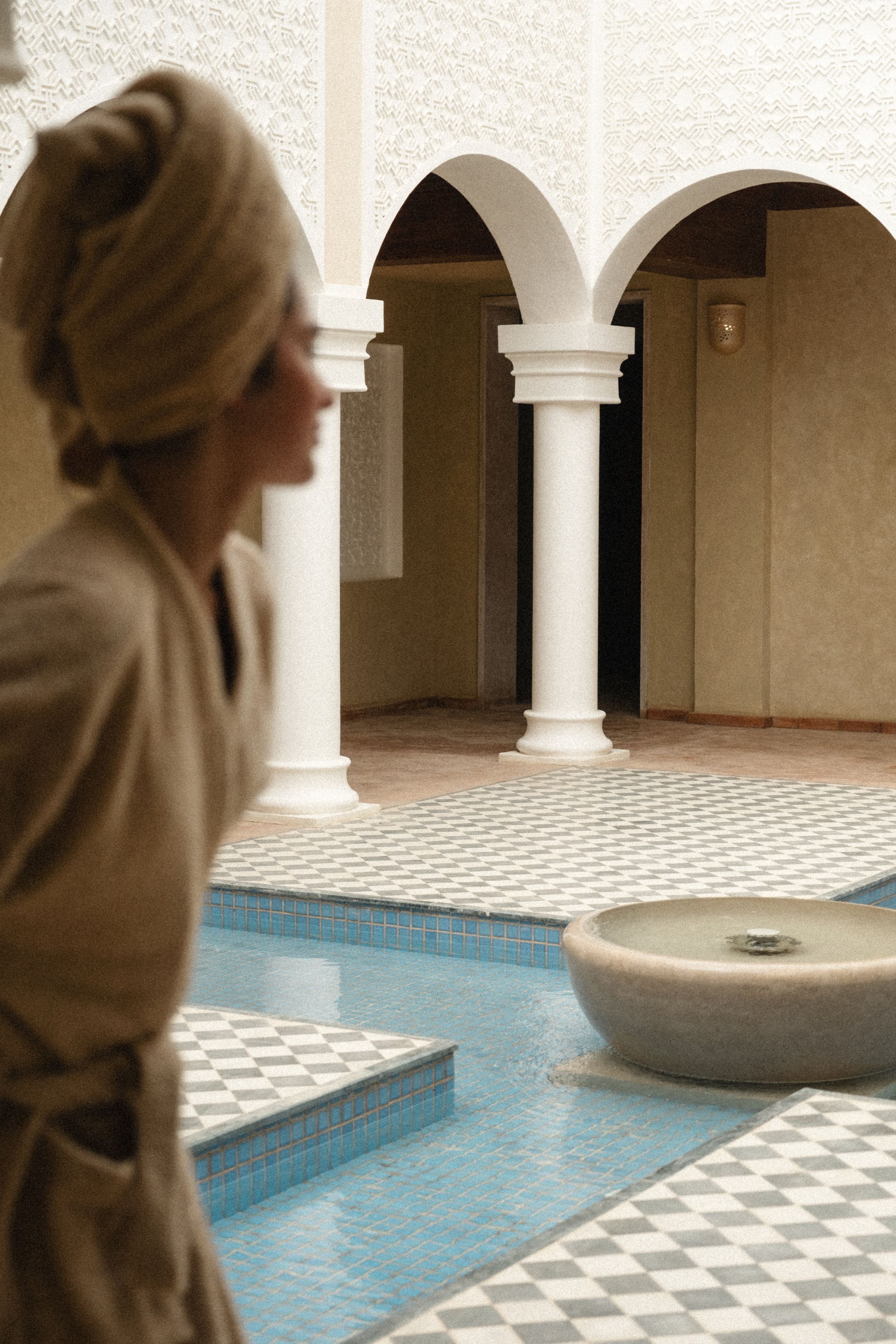A person in a robe walking through a courtyard with a pool and tiled flooring.