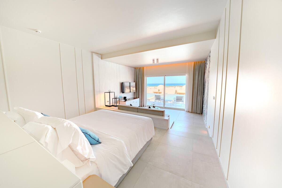 A modern hotel room with a large bed, a desk with a chair, and a view of the beach through the window.