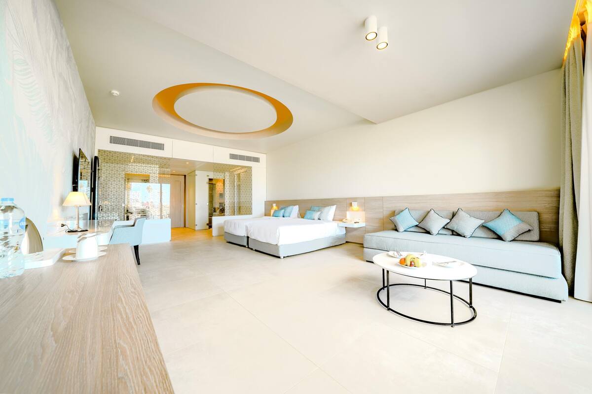 A modern hotel room with a large bed, a sofa, a coffee table, and a desk with a lamp.