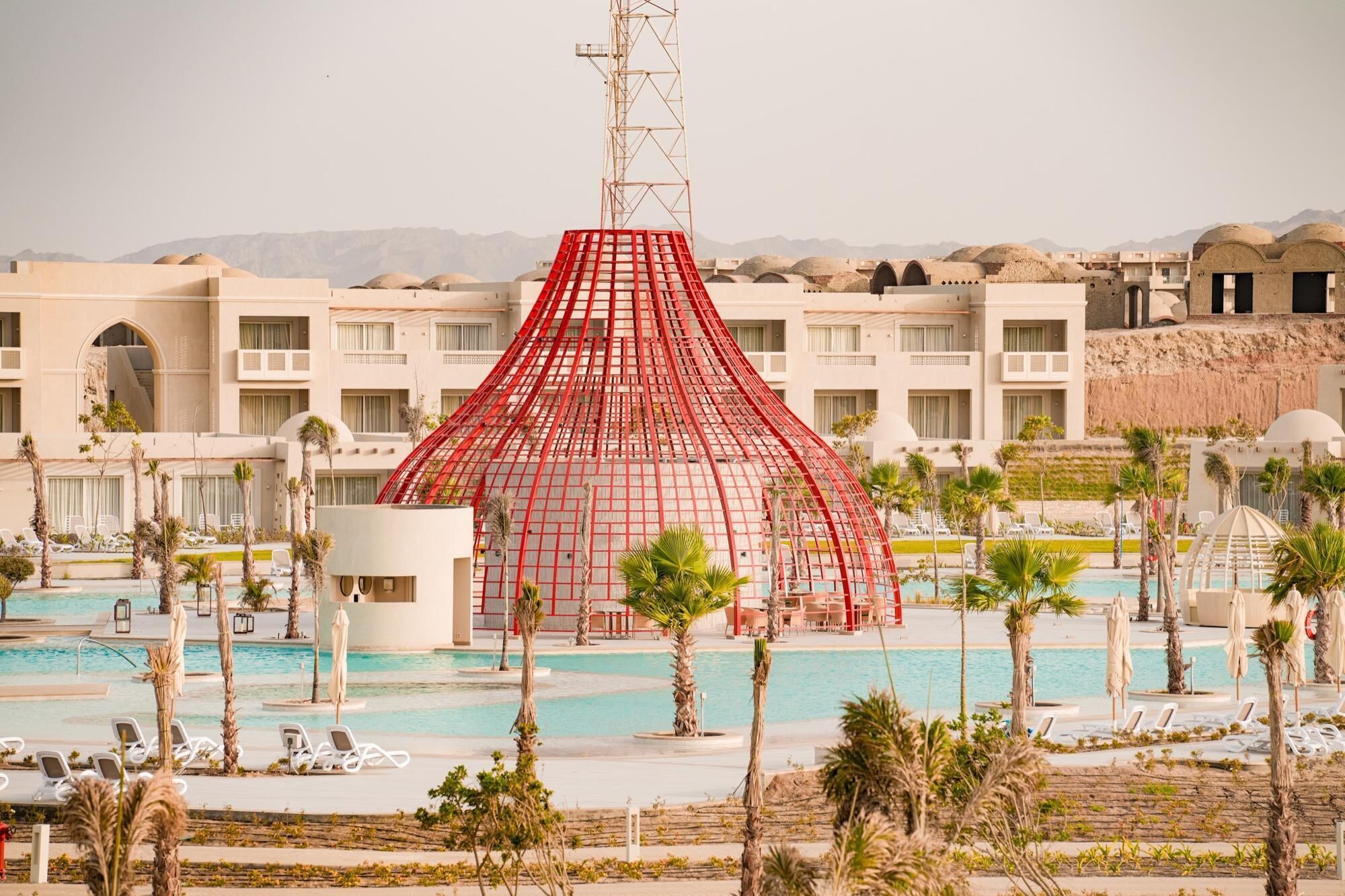 A hotel with a large pool, palm trees, and a distinctive red structure in the center.