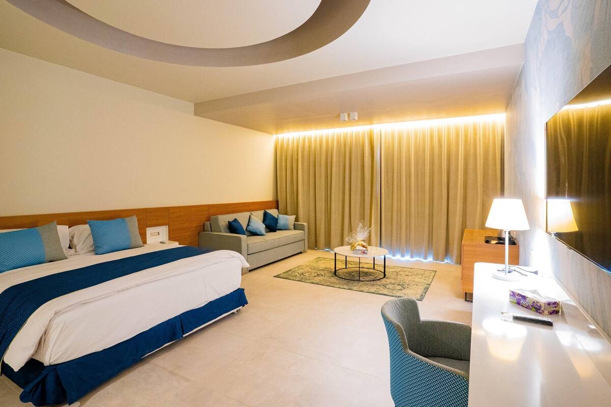 A hotel room with a large bed, a sofa, a desk with a lamp, and a TV.
