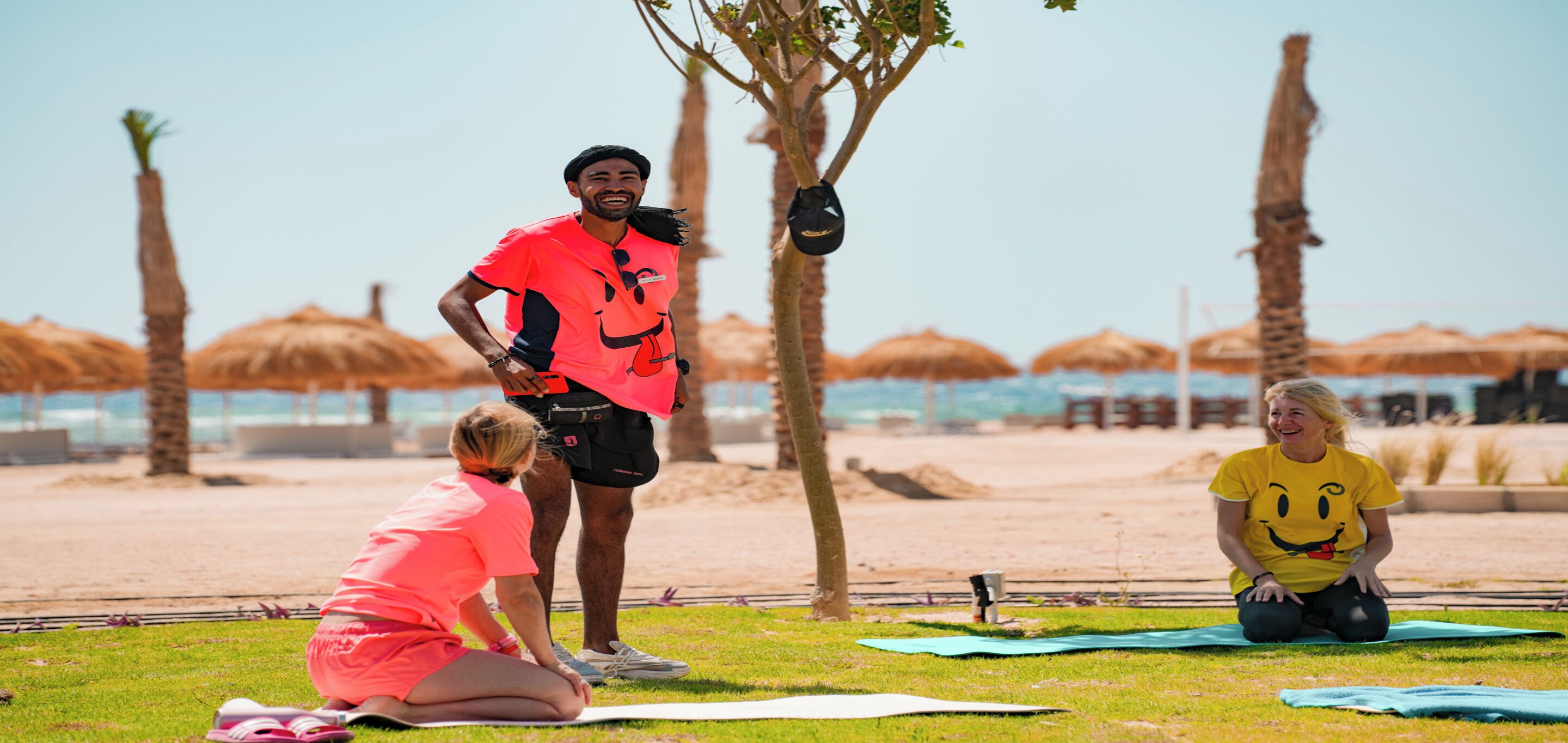 A beach setting with people on yoga mats, palm trees, and thatched umbrellas.