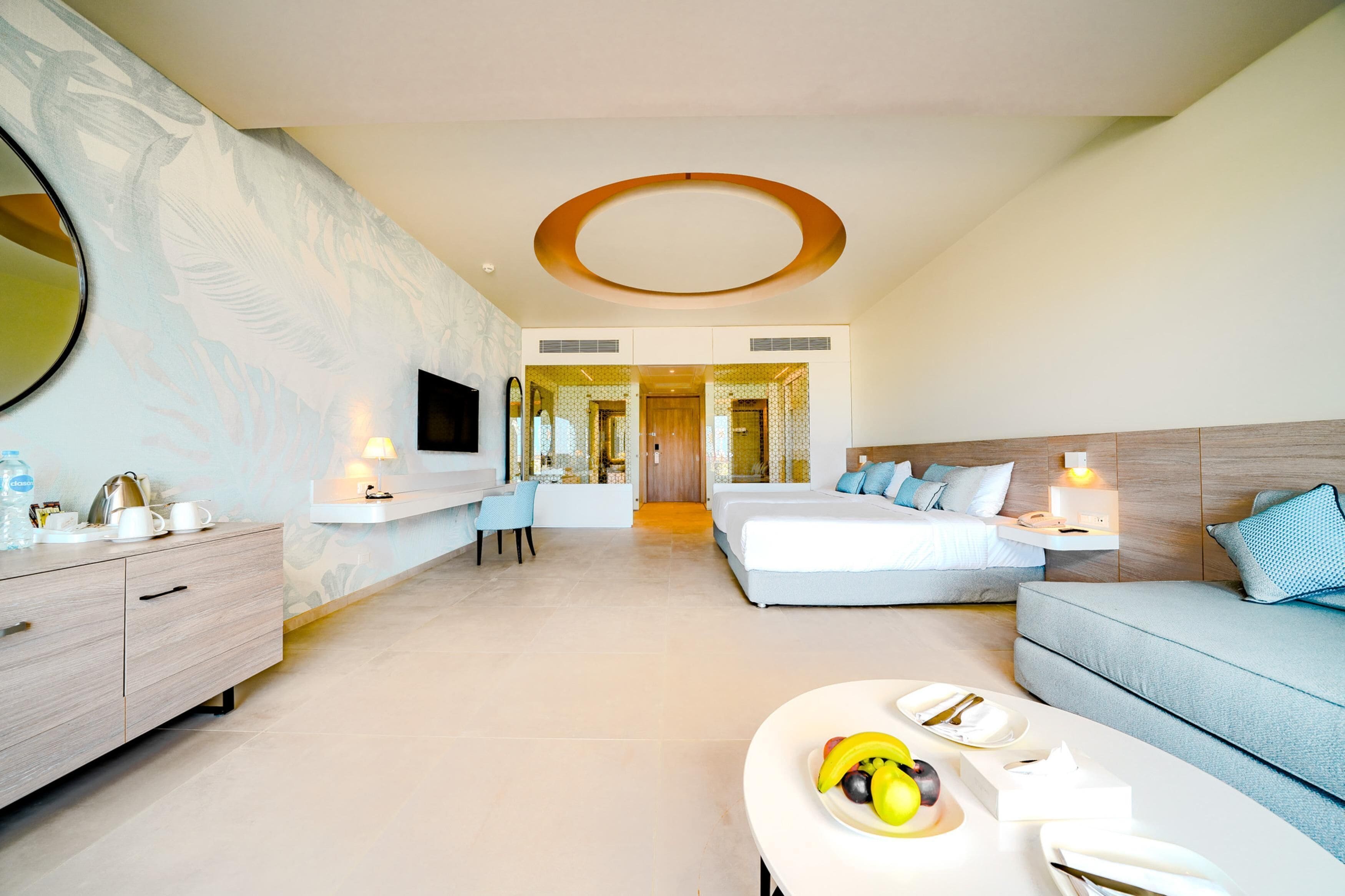 A modern hotel room with a large bed, a sofa, a dining area with fruit, and a TV.