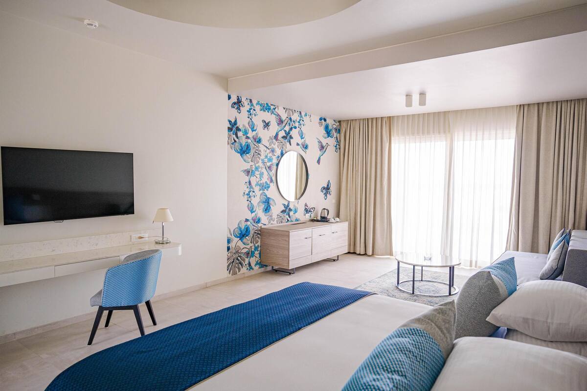 A modern hotel room with a large bed, a flat-screen TV, a desk with a chair, a vanity with a mirror, and a window with curtains.