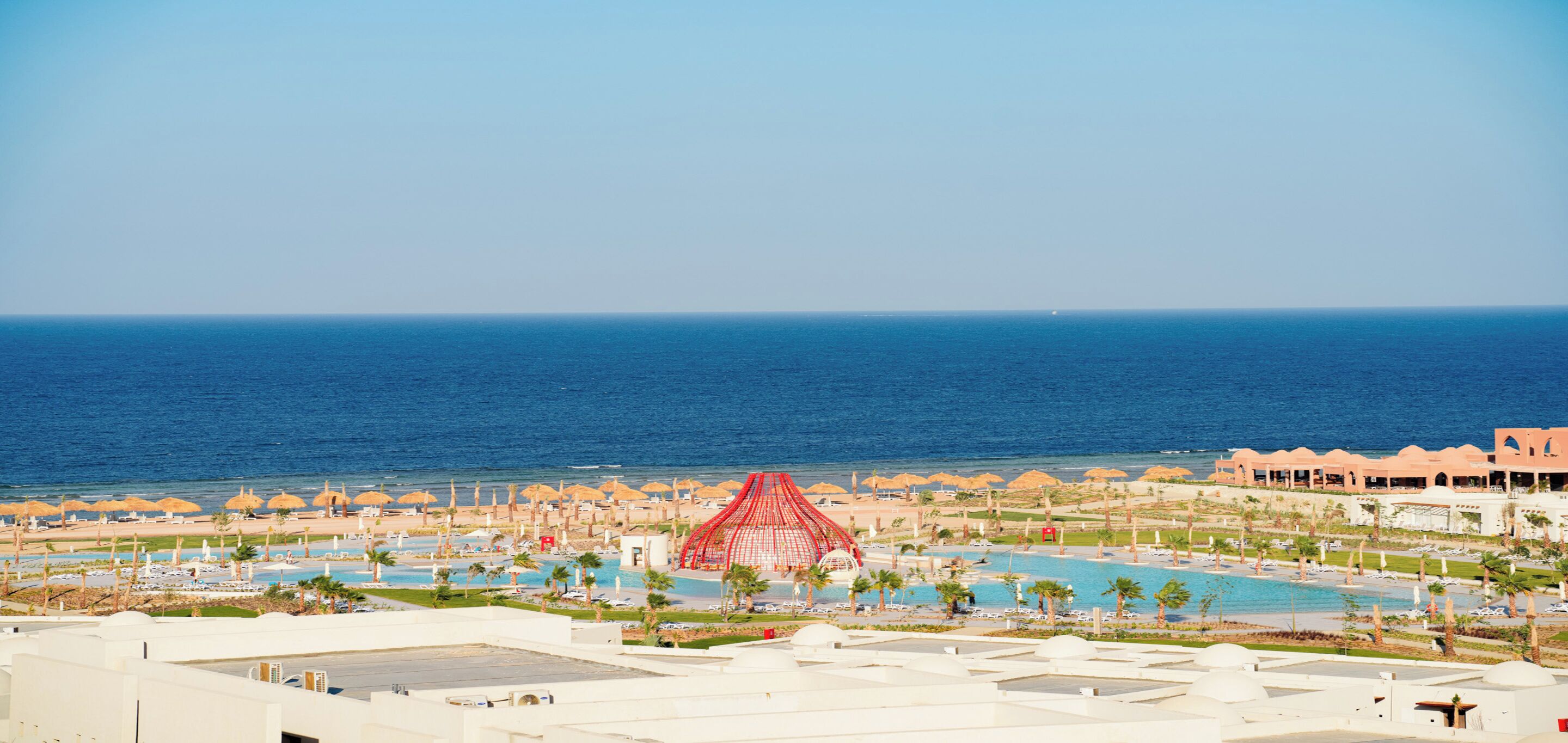 Un resort con diverse piscine, una grande struttura rossa e una spiaggia attrezzata con ombrelloni.