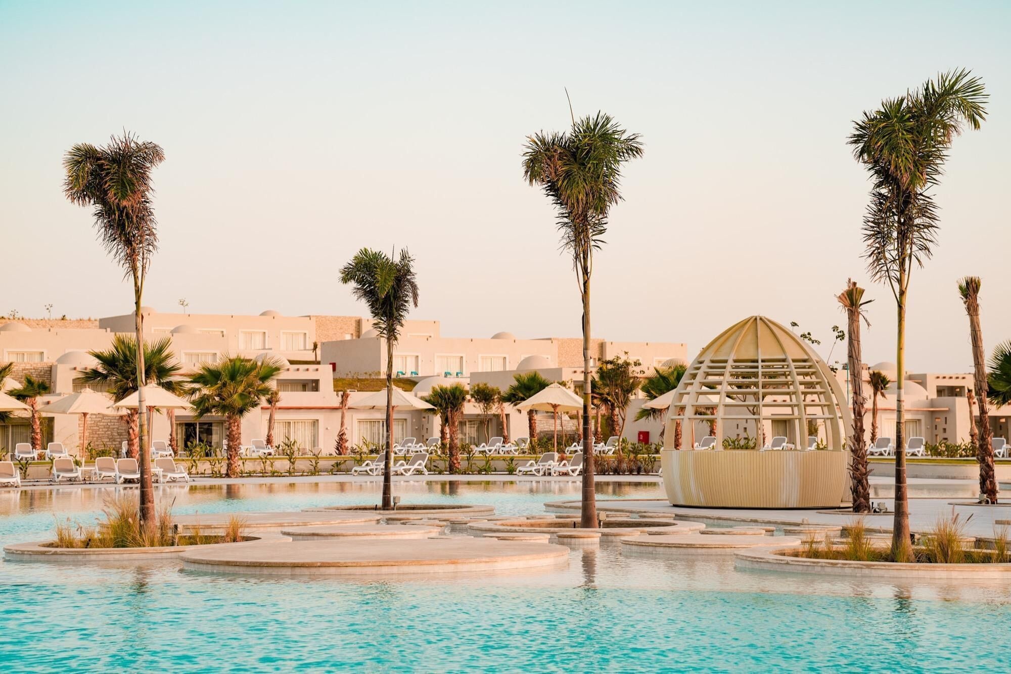 Zona de piscina de un resort con sillas de descanso, palmeras y una estructura de edificio singular.
