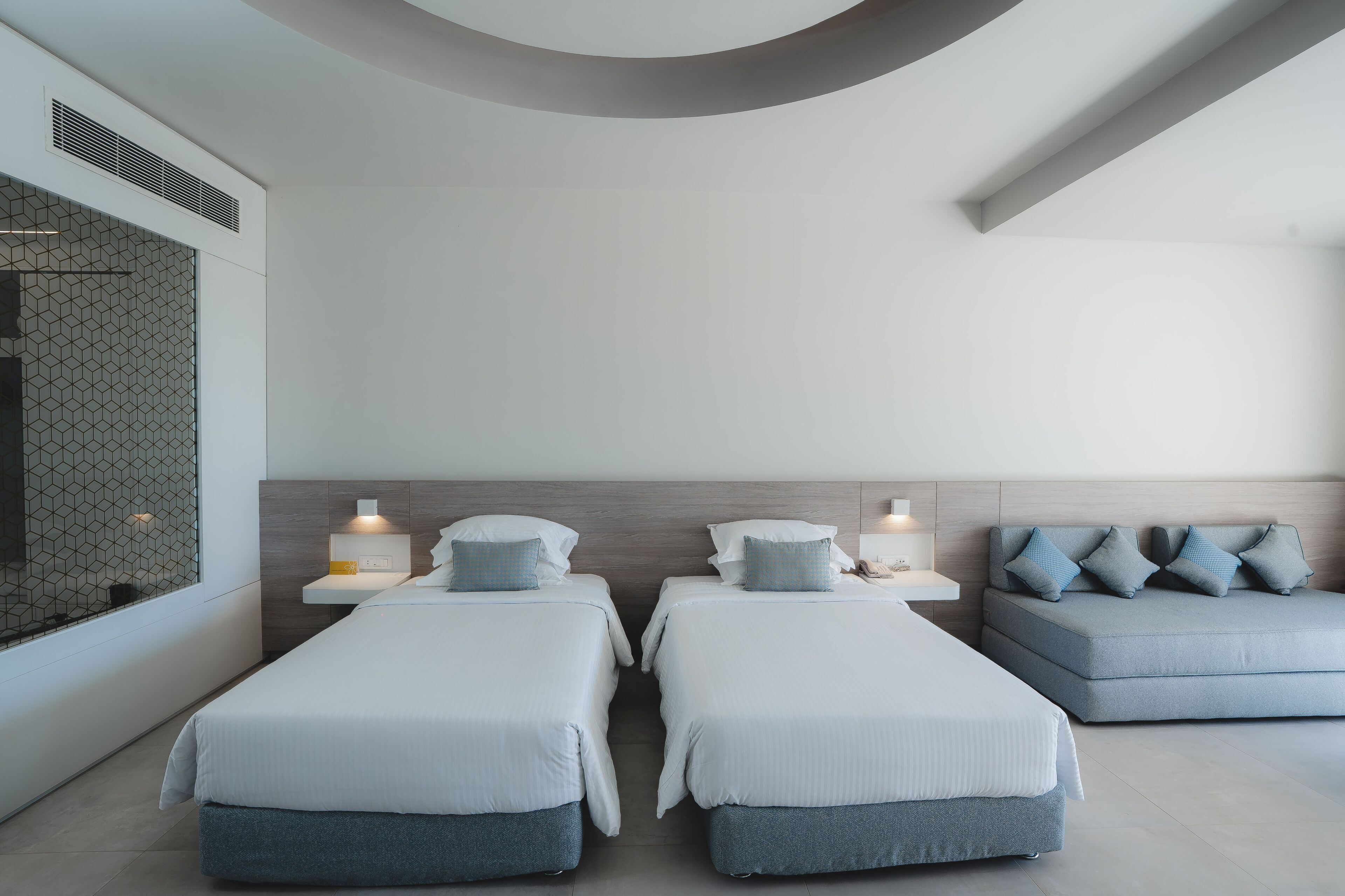 A modern hotel room with two single beds, a sofa, and a wall-mounted TV.