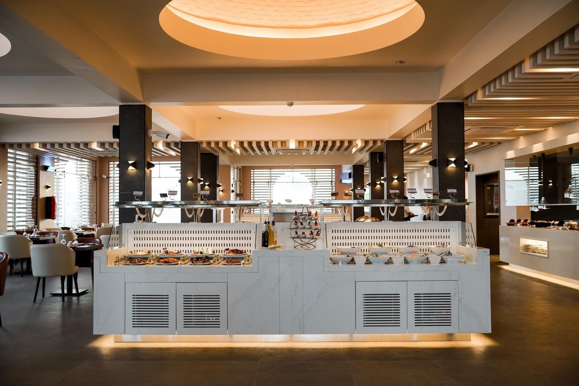 A modern restaurant with a central buffet area, multiple seating arrangements, and a contemporary ceiling design.