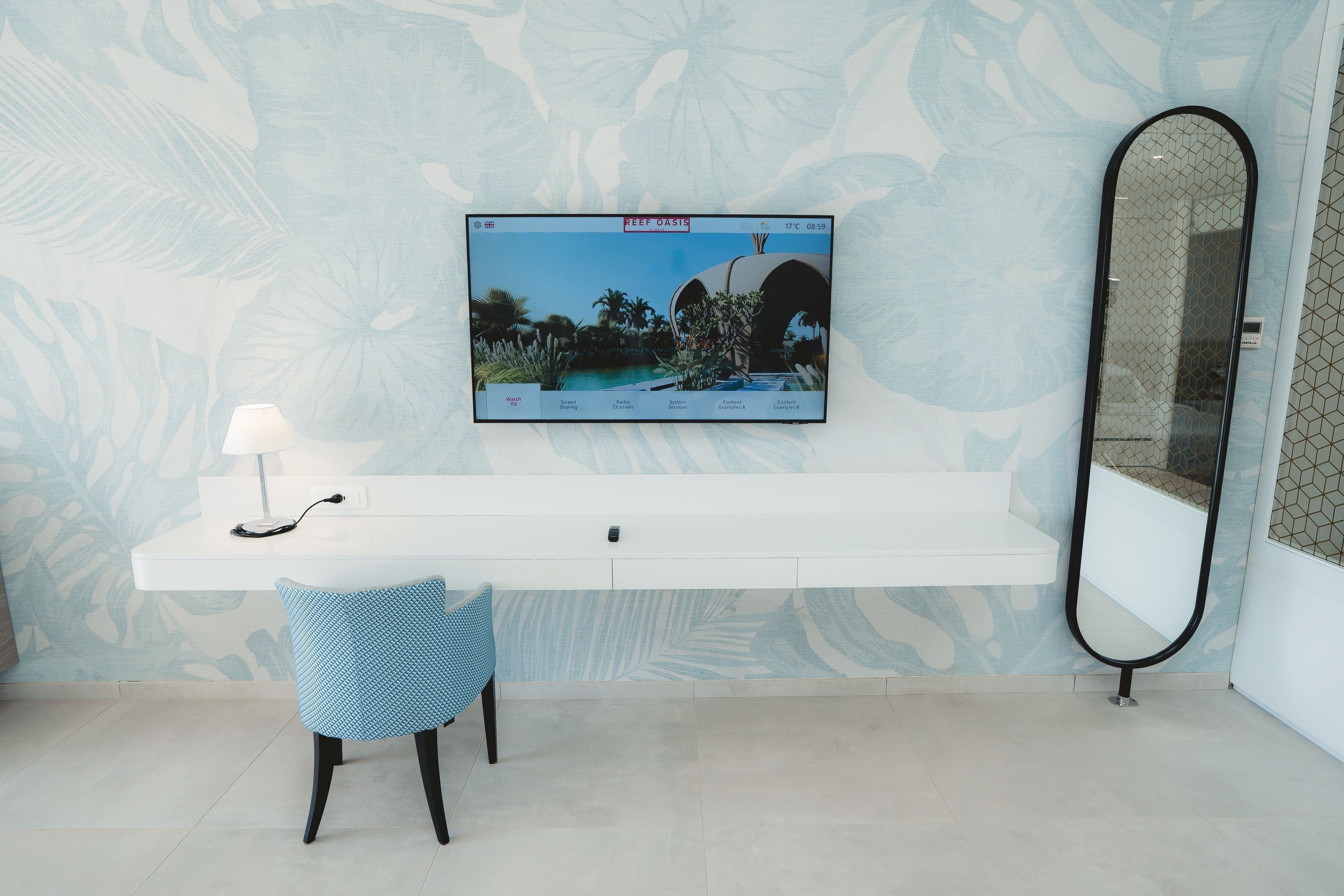 A modern bathroom with a large mirror, a mounted television displaying a tropical scene, a white vanity with a lamp, and a blue patterned chair.