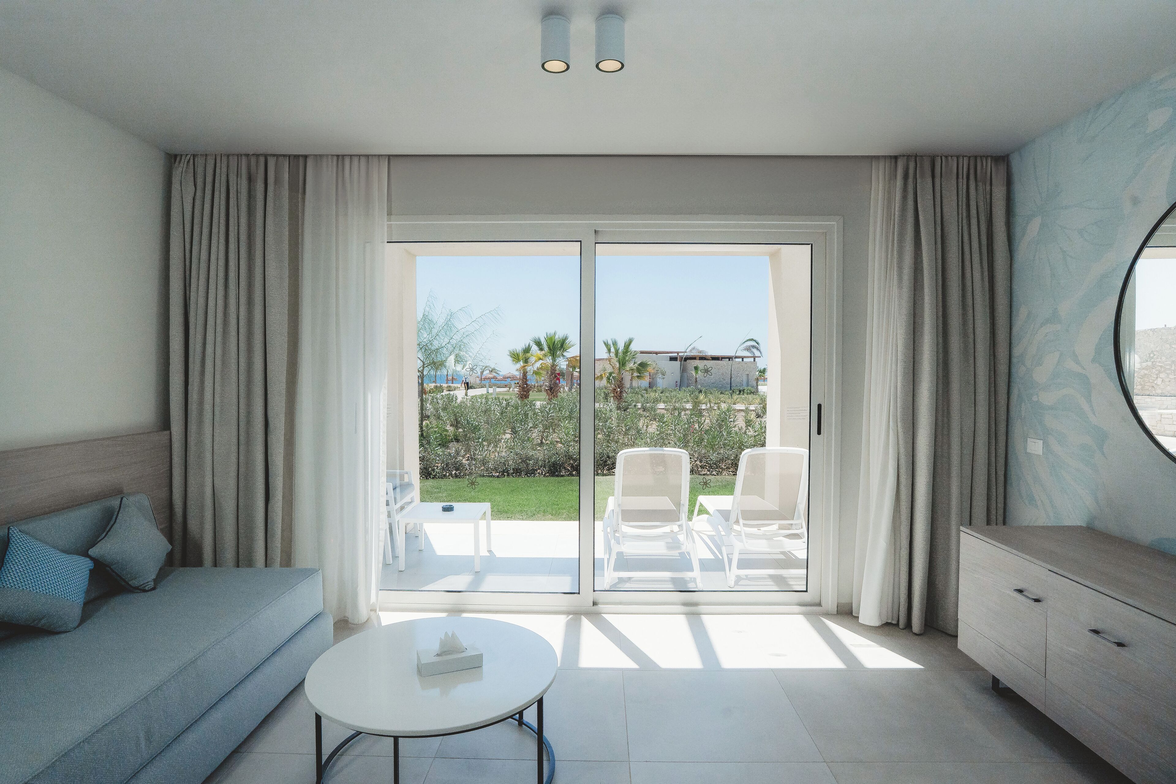 A modern hotel room with a sofa, a small table, a cabinet, and a sliding glass door leading to an outdoor seating area with chairs and a table.